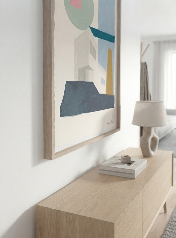 A framed  Modernist Architecture Wall Art featuring geometric house forms in a Mid-Century Modern composition by Francis Phylae for Phylae Posters, with geometric shapes in muted blue, teal, cream, and yellow tones hangs on a white wall above a light wood console table. The table holds a cream ceramic lamp and a stack of books in a softly lit modern interior.