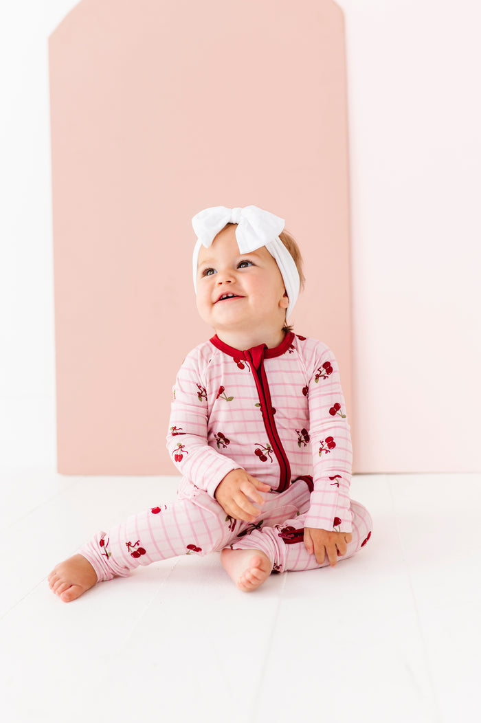 Baby wearing a pink outfit with red accents and a white headband, sitting against a light pink background.