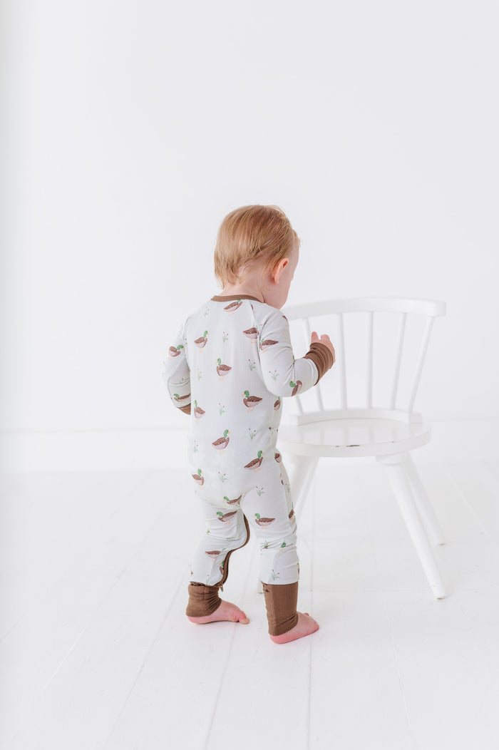 Child wearing a patterned onesie standing next to a white chair on a plain background